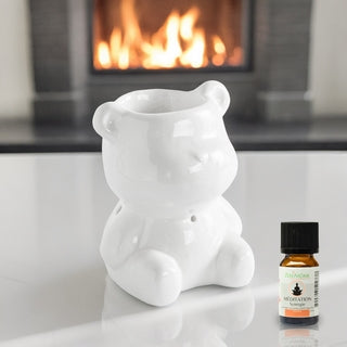 Brûle-parfum en céramique blanche en forme d'ourson modèle Teddy devant un feu de cheminée, avec un flacon d'huile essentielle, boutique KAVERN.