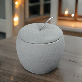 Brûle-parfum en céramique grise effet mat en forme de pomme, modèle Pomme d'Amour, sur une table en bois avec guirlande lumineuse en fond, boutique KAVERN.