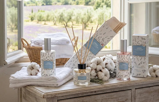 Gamme complète Fleur de Coton Le Chatelard 1802 présentée sur un meuble en bois devant une fenêtre ouverte sur les champs de lavande.