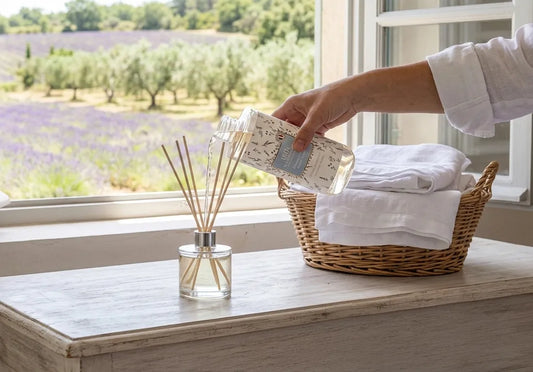 Remplissage d'un diffuseur de parfum à bâtonnets avec la recharge Fleur de Coton Le Chatelard 1802 devant une fenêtre ouverte sur la Provence.